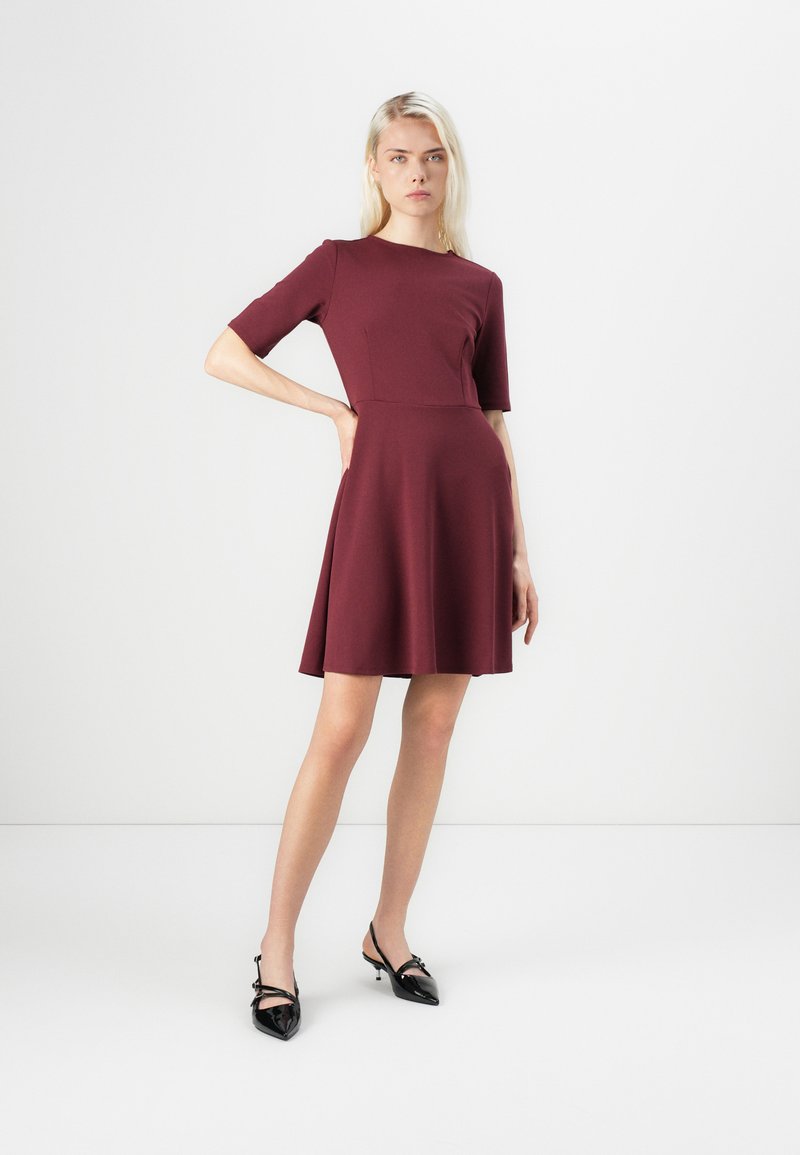 Burgunderfarbenes Kurzarmkleid mit tailliertem Oberteil und ausgestelltem Rock. Glattes Material mit einem dezenten Glanz. Schwarze spitze High Heels.