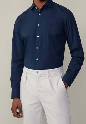 Homme portant une chemise bleu marine à manches longues boutonnée, rentrée dans un pantalon gris clair, avec une main dans la poche, sur un fond uni.