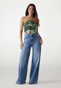 Grünes Neckholder-Top mit tropischem Blattmuster, kombiniert mit hellblauen Jeans mit weitem Bein. Glatte Stoffstruktur, lässige Passform, keine sichtbaren Verschlüsse.