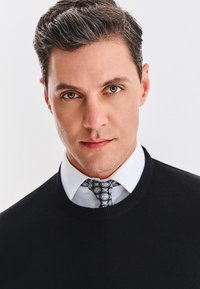 Czarny sweter z okrągłym dekoltem na białej koszuli z kołnierzykiem, wzorzysta krawat w odcieniach szaro-niebieskich, zbliżenie na twarz mężczyzny, neutralne tło.