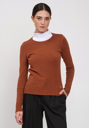 Vrouw draagt een roestkleurige trui over een witte coltrui, gecombineerd met losse zwarte broek, staand tegen een effen achtergrond.