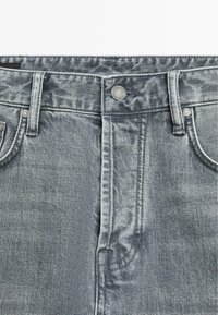 Le jean en denim gris présente un design classique à cinq poches, un lavage délavé et des accents en métal, incluant un bouton et des rivets.