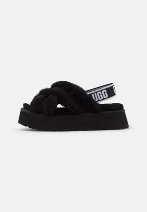 Sort platformsandal med tykke krydsede pelsede stropper og elastisk bagrem med mærke, der lyder "UGG" på hvid baggrund.