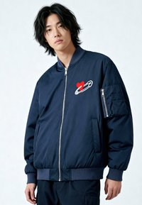 Giubbotto bomber blu navy con cerniera frontale, polsini a costine e ricami a forma di cuore e spilla da balia sul petto. Tessuto liscio e vestibilità rilassata.