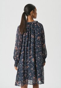 Vrouw met een donkere paardenstaart, gekleed in een marineblauwe bloemenjurk met lange mouwen en een gerimpelde hoge hals, met de rug naar de camera gekeerd tegen een effen achtergrond.