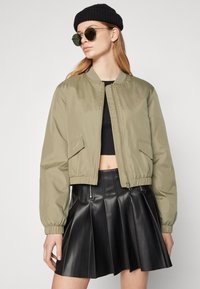 Olívovo zelená bomber bunda s elastickými rukávmi a pásom, so zipsom a bočnými vreckami, nosená cez čierny crop top a plisovanú koženú sukňu.