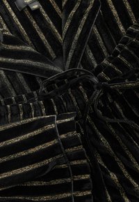 Juodas aksominis komplektas su auksinėmis metalinėmis juostelėmis. Audinys turi švelnią tekstūrą, su juosmens diržu ir klostuotais akcentais ant kelnių.