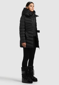 Čierna bunda typu puffer s kapucňou, vertikálne prešívaný dizajn a bočný popruh. V kombinácii s čiernymi legínami a textúrovanými zimnými boots.