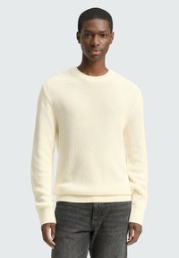 Lichtcreme gebreide trui met ribbelstructuur, ronde halslijn en lange mouwen, gecombineerd met donkergrijze jeans. Eenvoudig, tijdloos ontwerp.