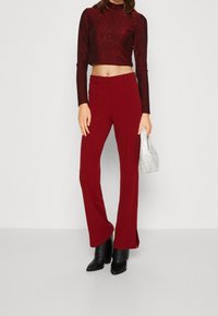 Rote, ausgestellte Hosen kombiniert mit einem glitzernden, kurzärmeligen Oberteil. Das Model hält eine silberne, metallische Handtasche. Schwarze Stiefel mit Absatz runden den Look ab.