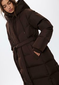 Smeđi zimski kaput s kapuljačom, remenom i velikim džepovima. Ima kvadratne sekcije i žnjanine, izrađen je od glatkog, sjajnog materijala.