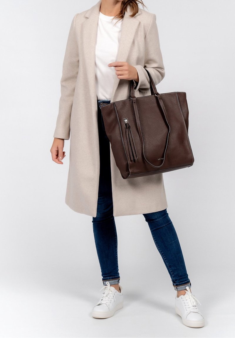 Personne portant un manteau beige, une chemise blanche, un jean bleu et des baskets blanches, tenant un grand sac cabas en cuir marron foncé par ses anses.
