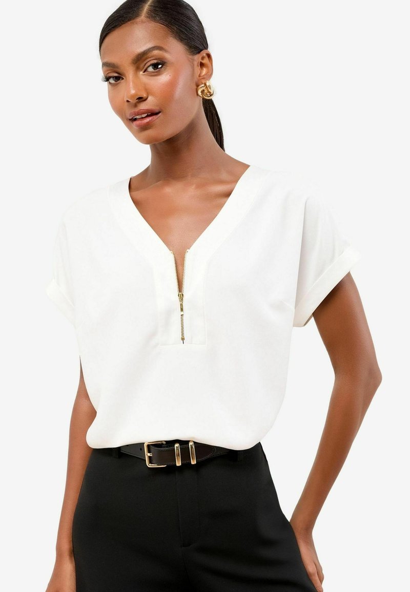White short-sleeve top with a V-neck and a central zipper. Paired with black pants and a black belt featuring a gold double buckle.
