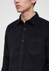 Jeune personne portant une chemise noire en velours côtelé à boutons avec une poche poitrine sur un fond clair uni.