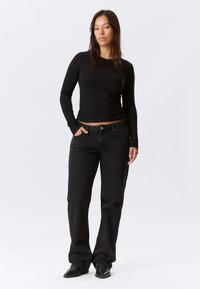 Top nero a maniche lunghe con design aderente, abbinato a jeans neri larghi. Il modello sta in piedi con le mani nelle tasche, indossando scarpe nere.