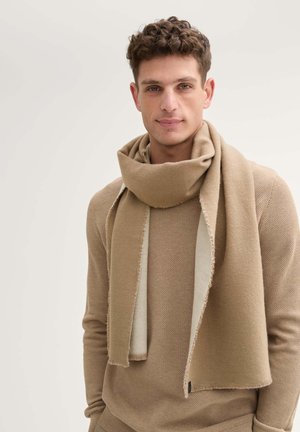 Bufanda de punto beige con una textura suave, anudada de forma suelta alrededor del cuello, con un interior beige más claro y bordes sin rematar, llevada con un suéter a juego.