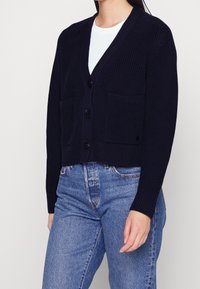 Marinblå stickad cardigan med V-ringning, knappdown fram och två fickor. Korthornad design med ribbad textur och långa ärmar.