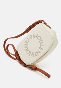 Kreemivärvi nahast kott, ümmargune kuju, pruun punutud rihm ja perforeritud logo kujundus esiküljel. Sisaldab reguleeritavat rihma.