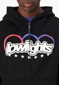 Sweat à capuche noir zippé avec un grand logo blanc agrémenté d'accents rouges, bleus et violets, ainsi que des étoiles blanches sur la partie inférieure.