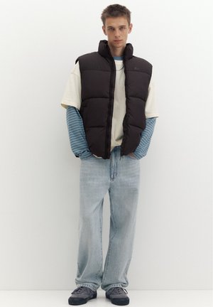 Jeune homme portant un gilet matelassé noir par-dessus des hauts à manches longues et courtes superposés, un jean bleu clair et des baskets grises, debout devant un fond blanc.