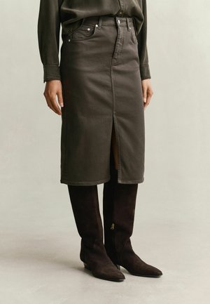 Personne portant une jupe foncée taille haute longueur genou avec une fente devant et des bottes foncées montantes avec fermetures éclair latérales.
