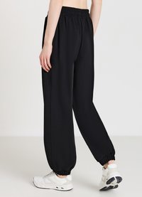 Pantalon de survêtement noir à coupe décontractée, avec une taille élastique et des poignets froncés. Porté avec des baskets blanches, mettant en valeur une texture lisse.