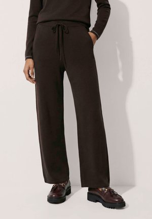 Person trägt schwarze Hose mit weitem Bein und Kordelzug in der Taille sowie dunkelbraune Loafers mit Quasten, steht vor hellem Hintergrund.