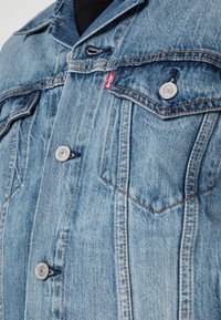 Světlomodrá džínová bunda s knoflíkovým zapínáním, dvěma kapsami na prsou a kontrastním prošitím. Na přední straně je malá červená značka značky.
