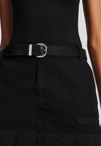 Svart skinnbelte med en sølvfarget, sirkelformet spenne, båret over et svart skjørt. Skjørtet har et brodert logo "KARL LAGERFELD JEANS."