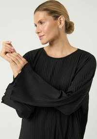 Svart plissert topp med lange voluminøse ermer, rund halsutringning og teksturert stoff. Har lett frynsete kanter på ermene.