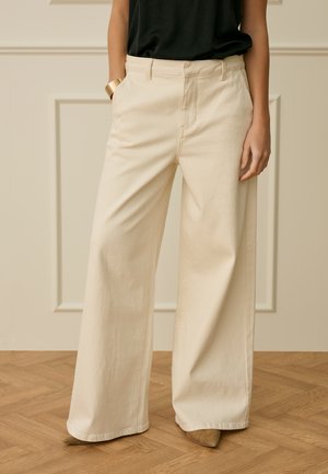 Kvinde iført beige bukser med vide ben og en sort top, stående på trægulv med hænderne i lommerne og en guldbøjle om håndleddet.