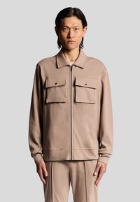 Beige zipjacka med långa ärmar, har två framfickor, en krage och en slät yta. Bärs med matchande byxor.