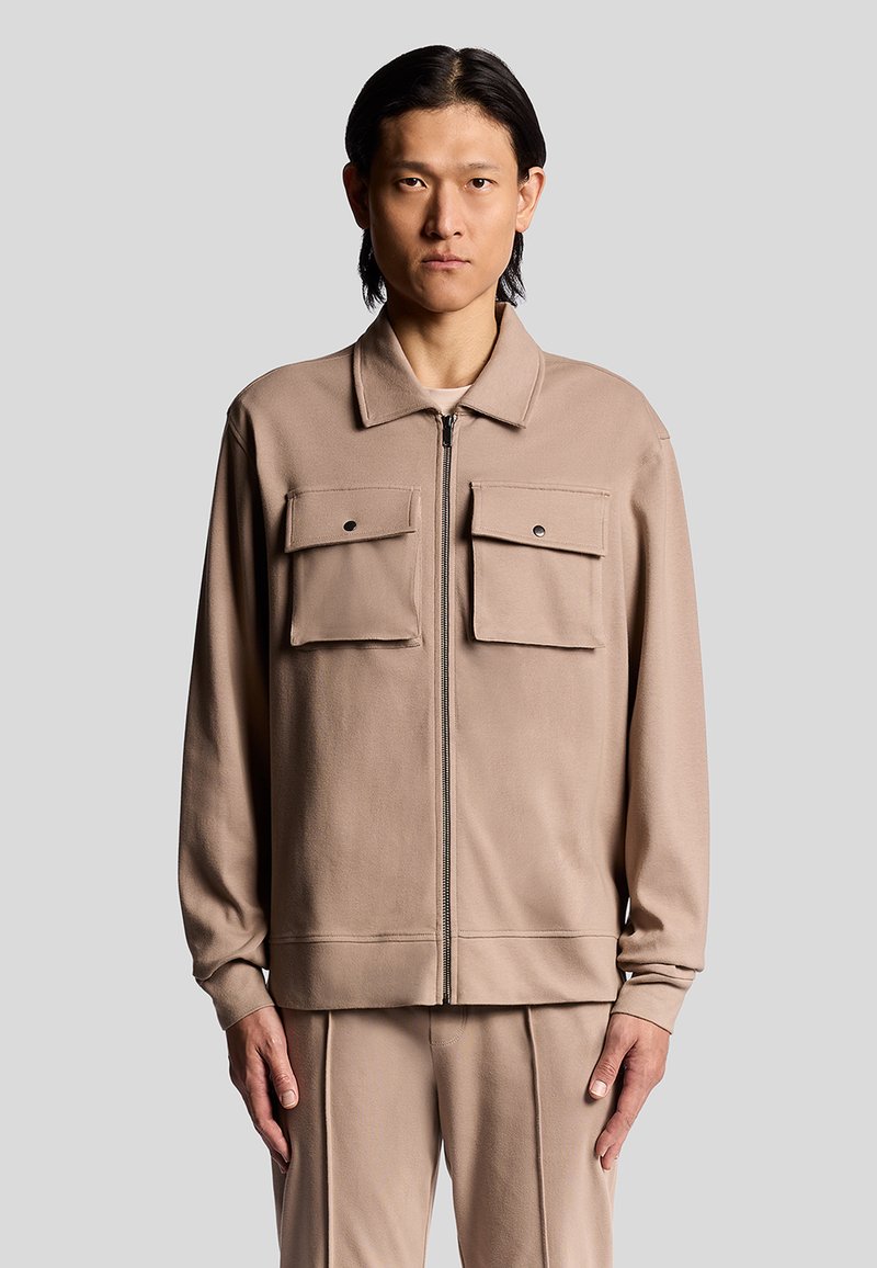 Beige zipjacka med långa ärmar, har två framfickor, en krage och en slät yta. Bärs med matchande byxor.