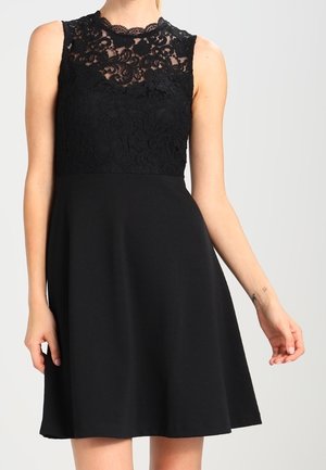 Czarna sukienka bez rękawów z koronkowym górnym panelem w kwiatowy wzór i jednolitą spódnicą w kształcie litery A, noszona przez osobę na jednolitym tle.