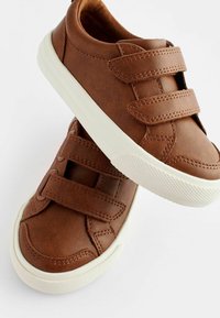 Brune syntetiske lædersko med to velcro-stropper, hvide gummisåler og struktureret dækmønster. Rund tådesign.