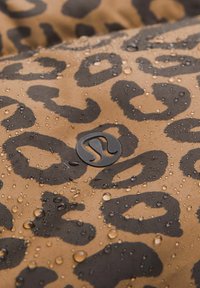 Voděodolná látka s hnědým pozadím, na kterém je vzor černých leopardích skvrn. Na povrchu jsou viditelné malé kapky. Přítomný detail loga.