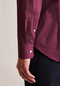 Vínová košile s bílým vzorem lístků, dlouhými rukávy a knoflíkovými manžetami. Hladká tkanina, přiléhavý střih a zakulacený lem.