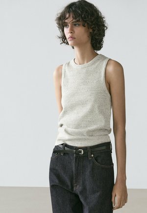 Jeune personne aux cheveux bouclés portant un haut sans manches gris clair et un jean noir avec une ceinture noire, debout devant un fond uni.