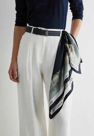 Person trägt weiße, hoch taillierte Hose mit einem schwarzen geflochtenen Gürtel und einem dunkel gemusterten Schal, der auf der rechten Seite in den Gürtel gesteckt ist.