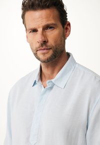 Světlomodrá lněná košile s límečkem, polo zapínáním a volnějším střihem. Materiál má hladkou texturu a jemný lesk.