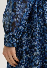 Blaue gemusterte Kleid mit unterschiedlichen Blautönen, schwarzen und weißen Punkten. Verfügt über einen gerafften Ärmel und eine geknöpfte Manschette.