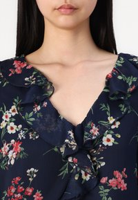 Blouse navy à motifs floraux avec un col à volants, présentant des motifs floraux blancs, roses et verts sur un tissu léger.