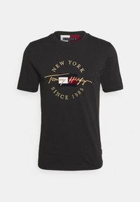 Černé tričko s krátkým rukávem a kulatým výstřihem, které má červené, bílé a modré logo a zlatý text "New York Since 1985 Tommy Hilfiger."