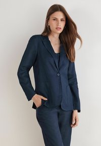 Marinblå linblazer med en enda knapp, spetsiga slag och framfickor. Slät textur, strukturerad design, kombinerad med en matchande topp.