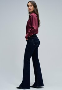 Mujer vistiendo una blusa de satén burdeos de manga larga y vaqueros acampanados oscuros, de pie con las manos en los bolsillos frente a un fondo liso.