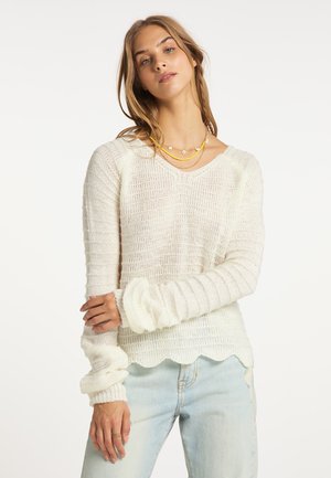 Junge Frau trägt einen weißen, strukturierten Strickpullover mit welligem Saum und hellblaue Jeans, steht mit sanft verschränkten Armen.