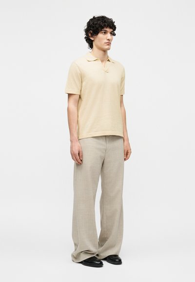 Νέος άνδρας στέκεται, φοράει μπεζ κοντομάνικη πόλο μπλούζα, φαρδιά ανοιχτογκρι παντελόνια και μαύρα παπούτσια σε απλό λευκό φόντο.