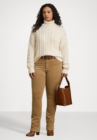 Krém színű, kábelmintás garbós pulóver, bézs kordbársony nadrág, krokodilbőr textúrájú barna kézitáska, barna magassarkú csizmák, álló póz.