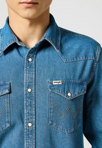 Camicia in denim blu medio con colletto a punta, bottoni a pressione, due tasche sul petto con motivo a zig-zag e etichetta del marchio Wrangler.