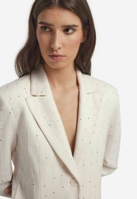 Biała marynarka z cekinowymi pionowymi paskami, głębokim dekoltem w kształcie V oraz wyraźnymi ramionami. Posiada jeden guzik i klapy z wycięciem.